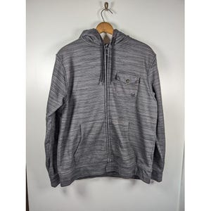May include: A gray zip-up hoodie with a horizontal striped pattern. The hoodie has a hood with drawstrings, a front pocket, and a small pocket on the chest. The hoodie is hanging on a wooden hanger.