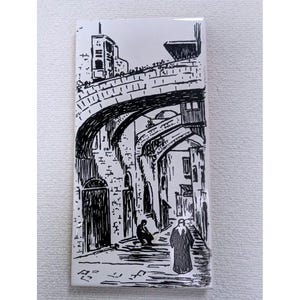 May include: A rectangular white ceramic tile featuring a black ink illustration of a narrow stone street scene with arched bridges, old buildings, and two figures walking along the path. The artwork displays detailed architectural lines and shadows.