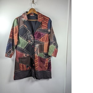 May include: A patchwork blazer in a variety of colors, including green, orange, and purple, hangs on a wooden hanger. The blazer features a collared neckline, two front pockets, and two buttons. The fabric appears to be a mix of textures and patterns.