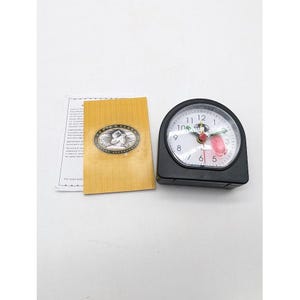 May include: A black analog travel alarm clock with a white face, black numerals, and a red second hand. Beside it lies a tan informational booklet featuring the Lewis N. Clark Travel Accessories logo, which depicts a person wearing a wide-brimmed hat.