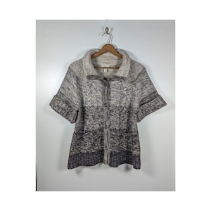 May include: A short-sleeved sweater in shades of gray, brown, and beige hangs on a wooden hanger. The sweater features a button-up front and a textured knit design. The collar is wide and the sleeves are cuffed.