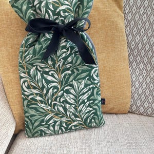 May include: A hot water bottle cover with a green and white botanical print, tied with a black ribbon. The cover is on a sofa with yellow and patterned cushions. The fabric design features leaves and stems.