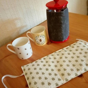 May include: A white ceramic mug with a bee pattern, a yellow ceramic mug with the text "Don't Worry Bee Happy", and a glass French press with a grey fabric cover. The French press has a red lid. All items are on a wooden surface.