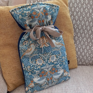 May include: A teal hot water bottle featuring a floral and bird design, secured with a beige ribbon. The pattern includes white flowers, orange berries, and birds. The bottle is on a yellow cushion.