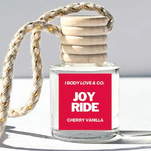 May include: A clear glass bottle with a wooden lid and a red label that says "I Body Love & Co. Joy Ride Cherry Vanilla". The bottle is hanging from a white rope with gold accents.