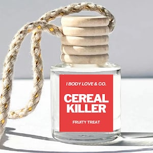 May include: A clear glass bottle with a red label that says "I Body Love & Co. Cereal Killer Fruity Treat" hanging from a white rope with gold accents. The bottle has a wooden cap with multiple layers.
