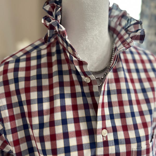 May include: A long-sleeved button-down shirt featuring a red, white, and navy blue plaid pattern. The shirt has a high, ruffled collar and a front button placket. A silver-toned chain necklace is visible around the neck of the mannequin.