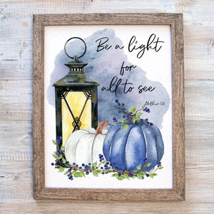 May include: A framed print with a watercolor painting of a lantern, two pumpkins, and a leafy vine with berries. The text "Be a light for all to see" is written in black script with the scripture reference "Matthew 5:16" below it.