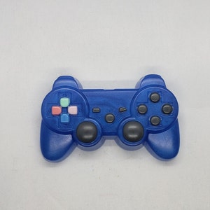 Pode incluir: Um controlador de videogame de plástico azul com botões pretos e um direcional de quatro vias. O controlador tem um botão azul e rosa no centro.