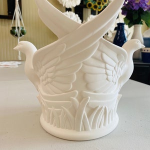May include: White ceramic vase with two doves in flight, surrounded by floral and grass designs. The vase is a blank canvas for painting.