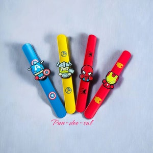 May include: Four colourful plastic wristbands featuring cartoon superhero characters: Captain America, Thor, Spiderman, and Iron Man. The wristbands are arranged in a fan shape on a white textured surface. The text "Pun-dee-sal" is written in pink below the wristbands.