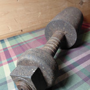 May include: A weathered, industrial-style metal object with a cylindrical body, a threaded central shaft, and a square-shaped end. The object is dark gray with signs of age and wear, suggesting it is vintage or antique.