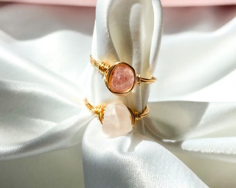 May include: Two gold rings with pink rose quartz stones wrapped in wire. The rings are displayed on a white satin fabric.