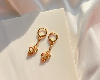 May include: A pair of gold hoop earrings with a snake charm dangling from each hoop. The snake charms are adorned with small clear rhinestones.