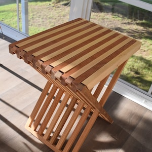 May include: A wooden folding stool with a striped top made of light and dark wood. The stool is made of natural wood and has a simple, rustic design.