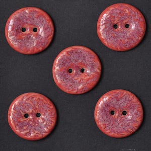 May include: A set of three ceramic mugs with a speckled, cream-colored glaze. Each mug features a different hand-painted design: a blue and green leaf, a yellow flower, and a red berry. The mugs have a simple, rounded shape.