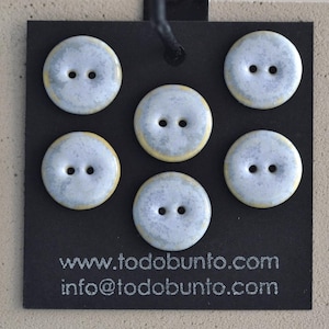 Puede incluir: Cinco botones redondos de dos agujeros con un acabado moteado en azul claro y gris y bordes amarillos, dispuestos sobre una tarjeta de presentación negra. La tarjeta incluye el texto www.todobunto.com e info@todobunto.com en blanco.