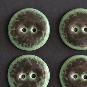 Könnte beinhalten: Vier runde Knöpfe mit einem Zwei-Loch-Design. Jeder Knopf hat eine glatte, glänzende Oberfläche mit einem gesprenkelten Farbmuster, das aus einem hellen mintgrünen Außenrand besteht, der in einen dunklen, bräunlich-schwarzen Mittelbereich übergeht.
