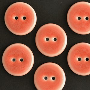 Puede incluir: Seis botones redondos de color coral con dos agujeros cada uno, dispuestos sobre un fondo negro. Los botones tienen un acabado brillante y un aspecto ligeramente moteado, lo que sugiere un material cerámico o esmaltado. Ideal para costura o manualidades.
