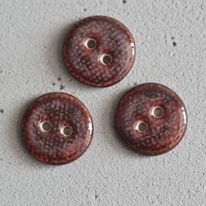 Op de afbeelding: Drie ronde, donkerbruine knopen met elk twee gaten. De knopen hebben een getextureerd oppervlak en een glanzende afwerking. De knopen zijn gerangschikt op een lichtgrijze ondergrond.