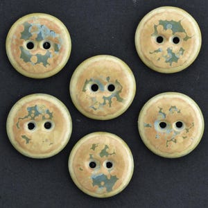 Puede incluir: Un conjunto de cinco botones redondos de dos agujeros con un patrón de esmalte moteado y variado en tonos beige, marrón claro y azul apagado. Cada botón tiene una superficie circular lisa con dos agujeros centrales para su fijación.