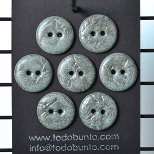 Puede incluir: Siete botones redondos de dos agujeros con una superficie moteada y texturizada en tonos gris y azul verdoso. Los botones están dispuestos sobre una tarjeta negra con el texto www.todobunto.com e info@todobunto.com impreso en blanco abajo.