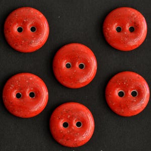May include: Six round, bright red buttons with two holes each. The buttons have a speckled texture and are arranged on a dark background. These buttons are suitable for sewing and craft projects.