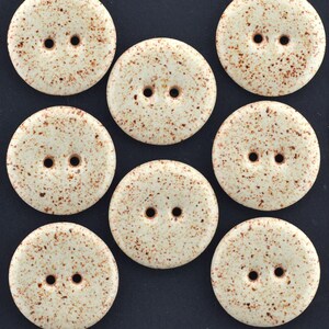 Puede incluir: Un conjunto de siete botones redondos de dos agujeros con una base de color crema y un patrón de motas marrones. Cada botón tiene una superficie circular lisa con dos agujeros centrados para su fijación, dispuestos en una cuadrícula sobre un fondo oscuro.