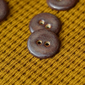 May include: Close-up of several brown ceramic buttons with two holes each, set against a textured mustard yellow fabric. The buttons have a glossy finish and a slightly irregular shape, suggesting a handmade quality. The fabric has a woven pattern.