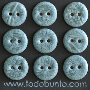 Puede incluir: Un conjunto de nueve botones redondos de color azul claro con un acabado texturizado y moteado, cada uno con dos agujeros centrales. Cada botón presenta patrones únicos, elevados y en espiral. El texto www.todobunto.com aparece abajo.
