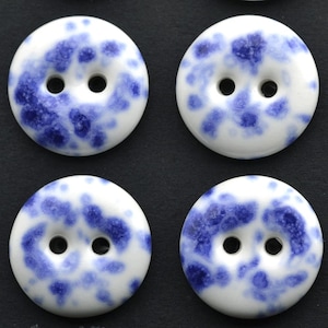 Puede incluir: Cuatro botones redondos con una base blanca y un patrón azul moteado. Cada botón cuenta con dos agujeros centrados para su fijación. La superficie de los botones parece lisa y brillante, mostrando un diseño distintivo de manchas azules y blancas.