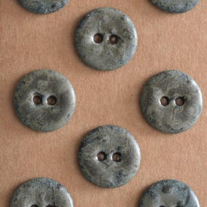 May include: A collection of round, two-hole buttons featuring a mottled, dark gray and charcoal textured surface. Each button has a smooth, slightly glossy finish with a distinct marbled appearance, arranged in a repeating pattern across the frame.