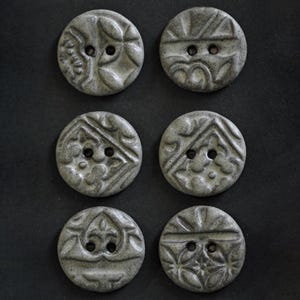 May include: A set of six circular gray ceramic buttons, each featuring a unique embossed pattern on the surface. Every button has two central holes for attachment. The buttons have a speckled, matte finish and are arranged in two vertical columns.