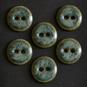 Puede incluir: Un conjunto de cinco botones redondos de dos agujeros con un esmalte brillante y moteado en tonos verde salvia, azul verdoso y marrón tierra. Cada botón tiene una superficie lisa ligeramente elevada con un acabado pulido.