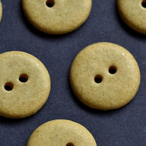 May include: Close-up of several round, light-yellow buttons with two holes each. The buttons are arranged on a dark surface. The buttons have a slightly textured appearance.