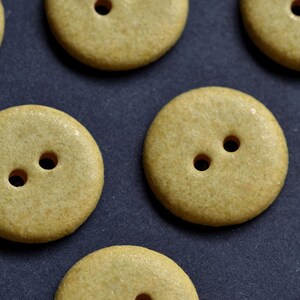 May include: A collection of round, tan-colored buttons, each featuring a smooth, speckled surface and two central holes for attachment. These circular fasteners are arranged in a pattern, showcasing their uniform shape and earthy, neutral tone.