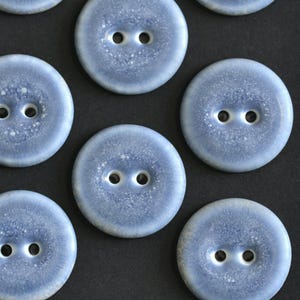 Puede incluir: Una colección de botones redondos de dos agujeros con un acabado moteado en azul claro y blanco. Cada botón tiene un centro liso y ligeramente cóncavo con un efecto degradado distintivo, que pasa de un borde azul oscuro a un centro más claro.