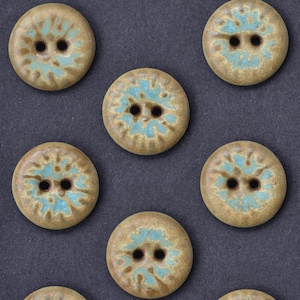 May include: A collection of round, ceramic buttons with a speckled, earthy brown base and a turquoise floral design. Each button has two holes for sewing. The buttons are arranged on a dark gray surface.