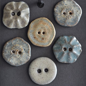 May include: A collection of six ceramic buttons in various shapes and colors. The buttons feature two holes for sewing and have a textured, glazed finish. Colors include shades of gray, blue, and brown. The buttons are arranged on a dark surface.