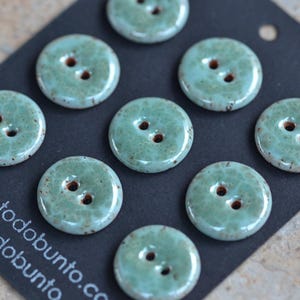 Puede incluir: Un conjunto de ocho botones redondos de dos agujeros con un esmalte brillante de color verde espuma de mar moteado. Los botones están dispuestos sobre una tarjeta negra con el texto blanco todobunto.com, parcialmente visible abajo a la izquierda.