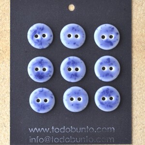May include: Nine round, pale blue buttons with a speckled, darker blue pattern. Each button has two holes for sewing. The buttons are displayed on a black card with the website address todobunto.com and an email address.