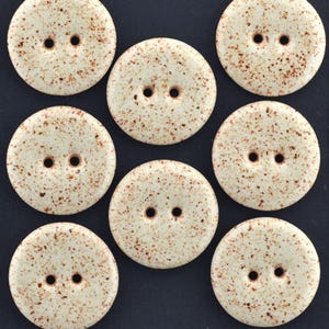 May include: A set of seven round, off-white buttons featuring a speckled brown pattern. Each button is circular with a smooth surface and contains two centered holes for attachment. The buttons are arranged in a grid-like pattern.