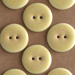 May include: Close-up of several round, pale yellow buttons with two holes each. The buttons have a speckled texture and a glossy finish, suggesting they are made of ceramic or a similar material. These buttons are suitable for sewing and craft projects.