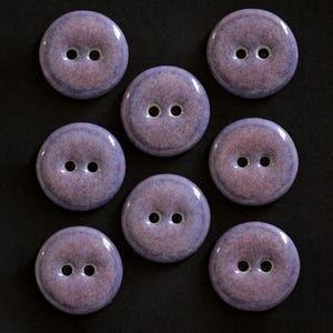 Puede incluir: Un conjunto de siete botones redondos de dos agujeros con un acabado brillante y moteado en tonos lavanda, morado y rosa suave. Cada botón tiene una superficie lisa y ligeramente abombada con un degradado de color hacia los bordes.