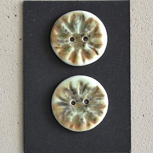 May include: Two round ceramic buttons with a floral design. The buttons are off-white with brown and green accents and two holes for sewing. They are displayed on a black background.