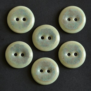 May include: A set of five round, two-hole buttons featuring a glossy, mottled glaze in shades of pale green, cream, and soft brown. Each button has a smooth, slightly concave centre and a raised outer rim, displaying a unique, variegated finish.