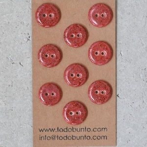 May include: A set of eight round, red buttons with two holes each, arranged on a brown card. The buttons have a speckled, glittery appearance. The card displays the website address www.todobunto.com and an email address.