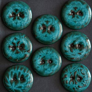 May include: A set of eight round buttons featuring a glossy teal and dark brown mottled glaze. Each button has two central holes and a smooth, reflective surface with unique color variations across the circular faces of the buttons.