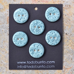 May include: Five round, light blue ceramic buttons featuring embossed snowflake patterns, each with two center holes. The buttons are attached to a black display card printed with the text www.todobunto.com and info@todobunto.com.