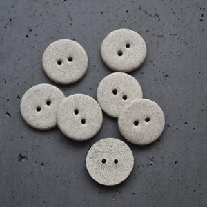 Puede incluir: Un conjunto de siete botones circulares de color gris claro con una textura moteada. Cada botón presenta dos orificios centrados para su fijación. Los botones tienen un acabado mate y una forma redonda, plana y uniforme.